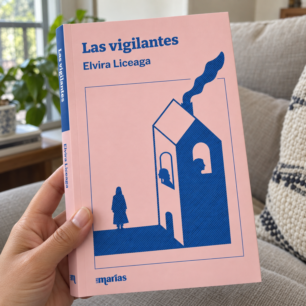 A hand holds a Spanish book titled 'Las vigilantes' by Elvira Liceaga. The book cover is light pink with a blue illustration of a house and a figure, and the publisher's name 'Las Marías' is visible at the bottom.